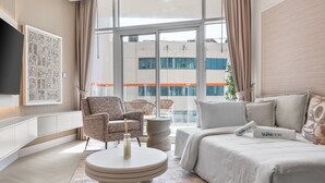 Una habitación de hotel moderna con una cama grande, una mesa de centro redonda, dos sillones, un sofá y vistas panorámicas de la ciudad a través de amplias ventanas.