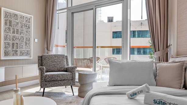 Habitación de hotel moderna con una cama grande, un área de descanso con una silla y una mesita, y ventanas amplias que ofrecen vistas a la ciudad.