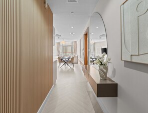 A modern hotel corridor with a wooden panel wall, a dining area with a table and chairs, and a vase with flowers on a counter.