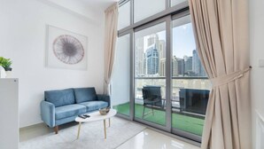 A modern living room with a blue sofa, a small white table, and a large window with curtains. - Classic Studio With Dubai Marina View (Dubai)