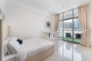 Une chambre moderne avec un grand lit, une Ă©tagĂšre intĂ©grĂ©e, une grande fenĂȘtre donnant sur des immeubles et un luminaire au plafond.