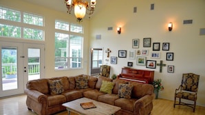 A spacious living room with a sectional sofa, a wooden coffee table, a piano, and a large window with a view of greenery. - Large home, perfect for family reunions (Ingram)