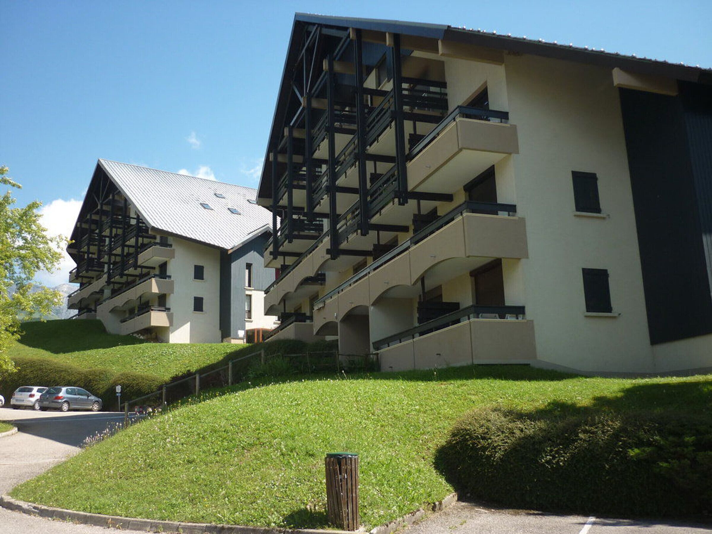 A modern multi-story building with balconies and a sloped roof, surrounded by well-maintained greenery.