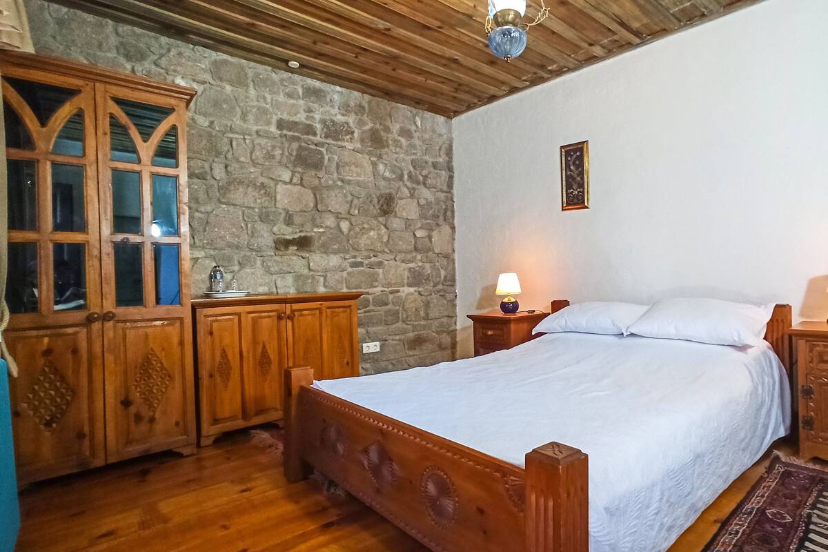 A bedroom with a wooden bed, two bedside tables with lamps, a stone wall, and a wooden wardrobe.