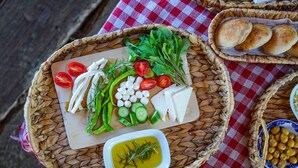 Uma mesa com toalha xadrez vermelha e branca, uma cesta com vegetais frescos, queijo e azeitonas, e uma pequena tigela de azeite.