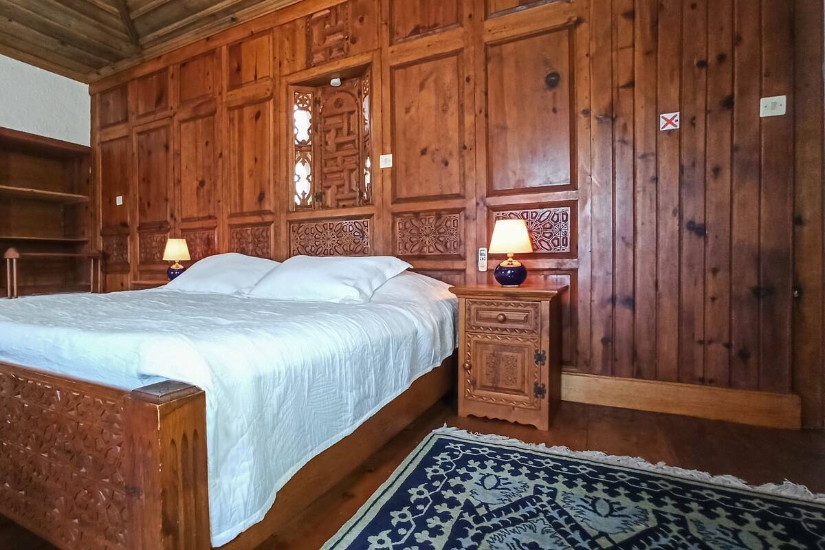 A wooden bedroom with a bed, two bedside tables with lamps, a nightstand, and a patterned rug.