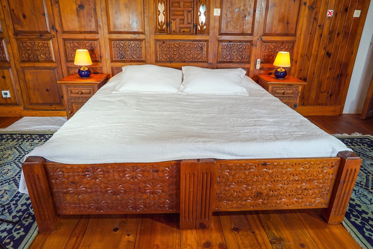 A wooden four-poster bed with a white bedspread, two nightstands with lamps, and a patterned rug on the floor.