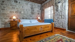 Uma cama de madeira com cabeceira, duas mesas de cabeceira com luminárias, uma parede de pedra, uma janela com cortinas e um tapete estampado.