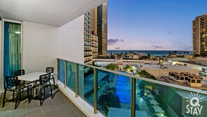 A balcony with a dining area, overlooking a pool and a cityscape with tall buildings and a view of the sea. - H'Residences – 2 Bedroom 1 Bathroom, Ocean View (Surfers Paradise)