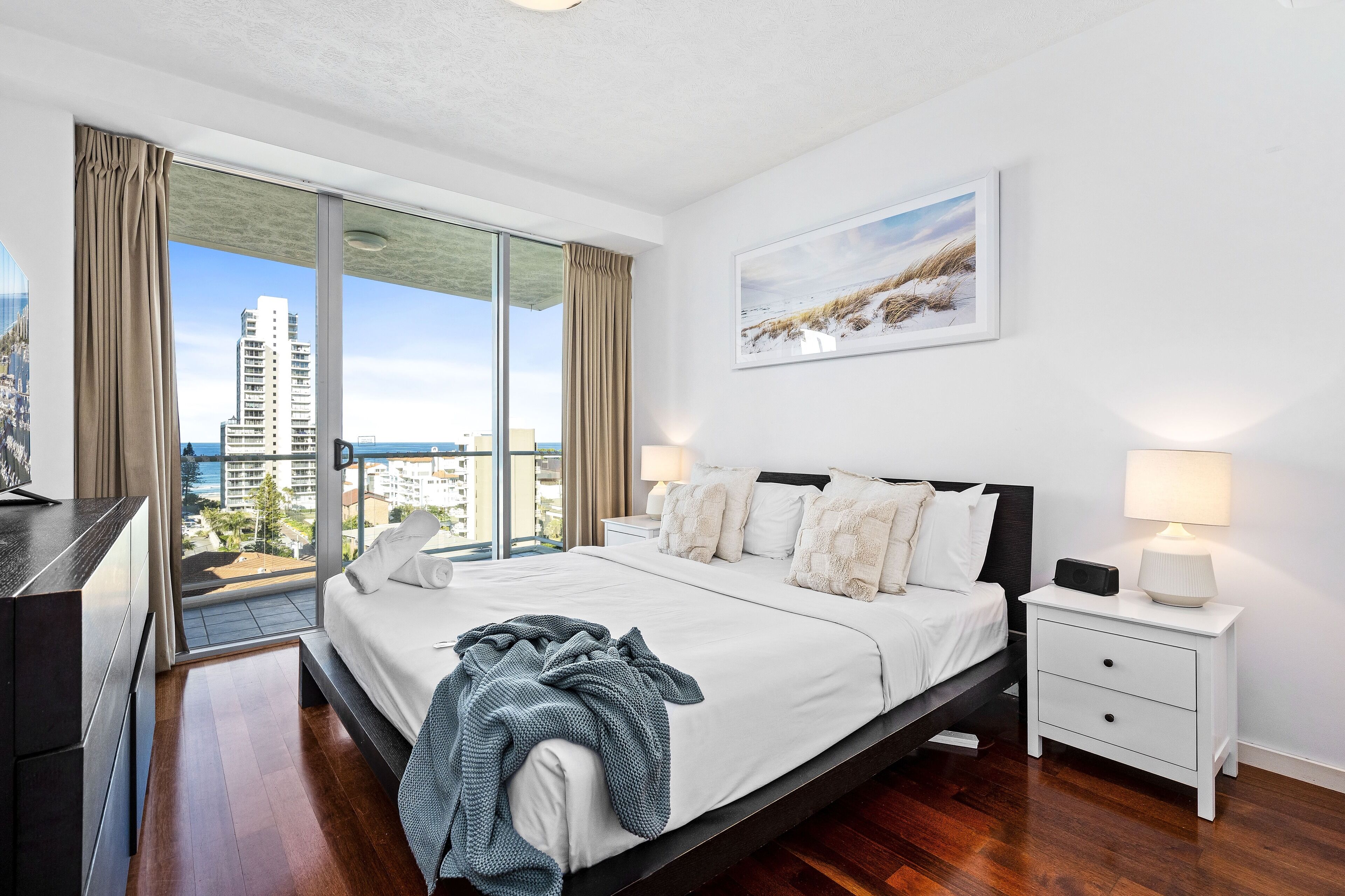 A modern bedroom with a large bed, a nightstand, and a view of the cityscape through floor-to-ceiling windows.
