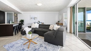 A modern living room with a dark grey sectional sofa, a glass coffee table, and a kitchen area with a dark countertop and bar stools. - 3 Bedroom Unit with Sea Views in Surfers Paradise (Surfers Paradise)