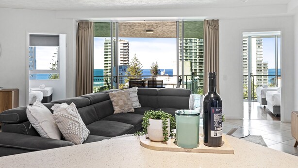 A modern living room with a dark grey sofa, a small table with a bottle and a plant, and a view of high-rise buildings and the sea through large windows. - 3 Bedroom Unit with Sea Views in Surfers Paradise (Surfers Paradise)