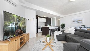 A modern living room with a large flat-screen TV, a grey sectional sofa, a glass coffee table, and a kitchen in the background. - 3 Bedroom Unit with Sea Views in Surfers Paradise (Surfers Paradise)