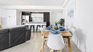 A modern kitchen with white cabinets, a central island, and a dining area with a wooden table and white chairs. - 3 Bedroom Unit with Sea Views in Surfers Paradise (Surfers Paradise)