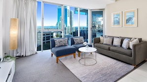 A modern living room with a grey sofa, blue armchair, and a small round table. Large windows offer a view of tall buildings and a cityscape. - H'Residences – Cozy 2 Bed Hinterland View — Q Stay (Surfers Paradise)