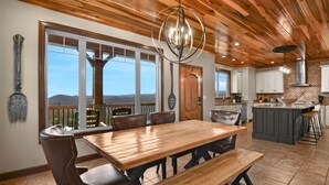 Un comedor amplio con una mesa de madera y bancos, una cocina con salpicadero de ladrillo y ventanas grandes que ofrecen vistas a las montañas.