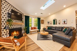 A modern living room with a fireplace, leather sofa, wooden bench, and a large TV. - Ski Slope Views Hot Tub & Best Resort Spot! (McGaheysville)