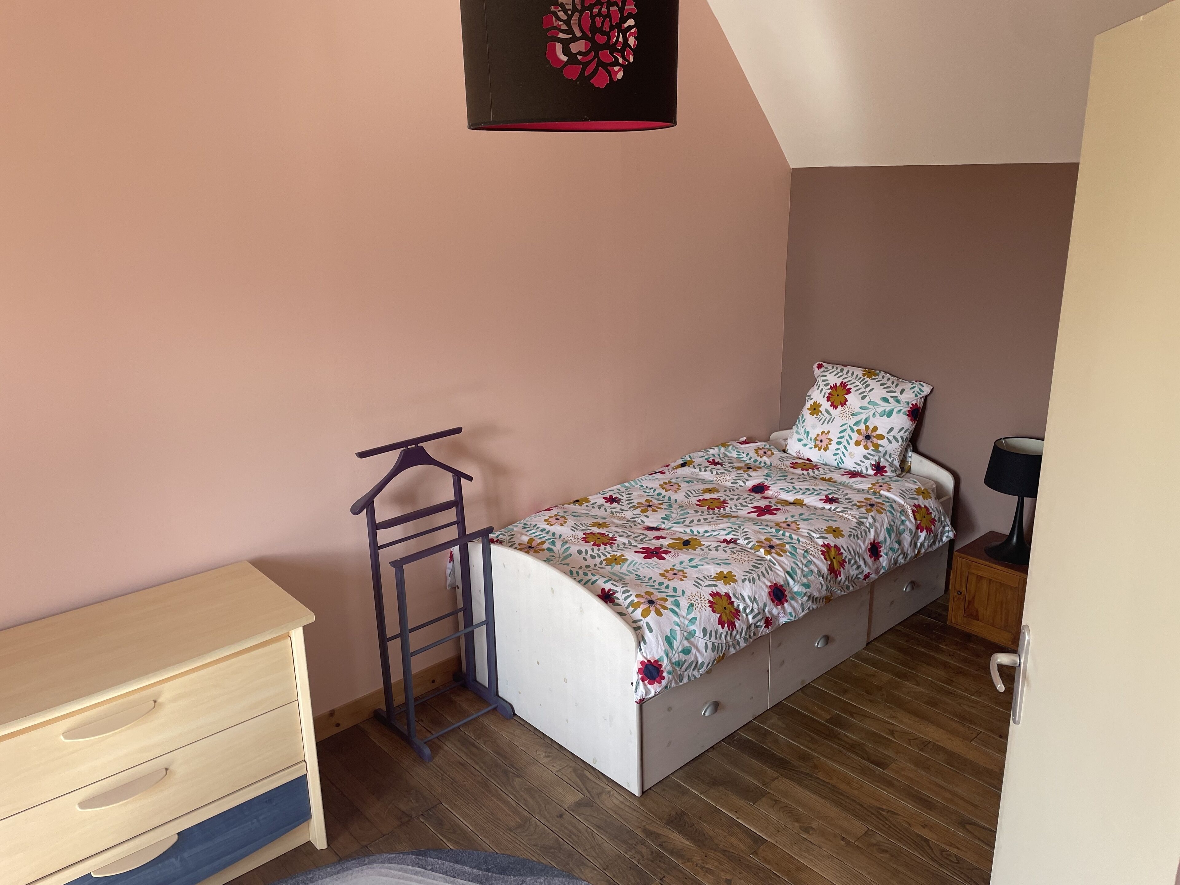 A small, single-bed room with a wooden dresser, a ladder, a desk, and a lamp.