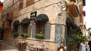 Un edificio de piedra con un restaurante llamado 'Gazetta Pizza & Pasta'. Hay mesas y sillas afuera, y se ve un reloj en la pared.