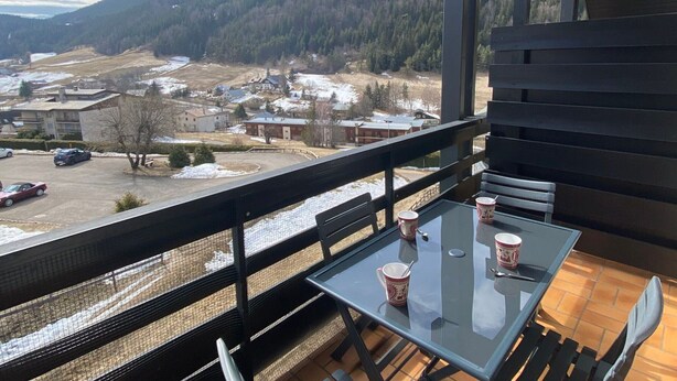 Un balcón con una mesa y sillas de vidrio, con vistas a un paisaje nevado con casas y una colina boscosa.