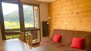 Una habitación con pared de madera, un sofá marrón con cojines rojos, una mesa de madera y vistas a un paisaje verde a través de un ventanal grande.