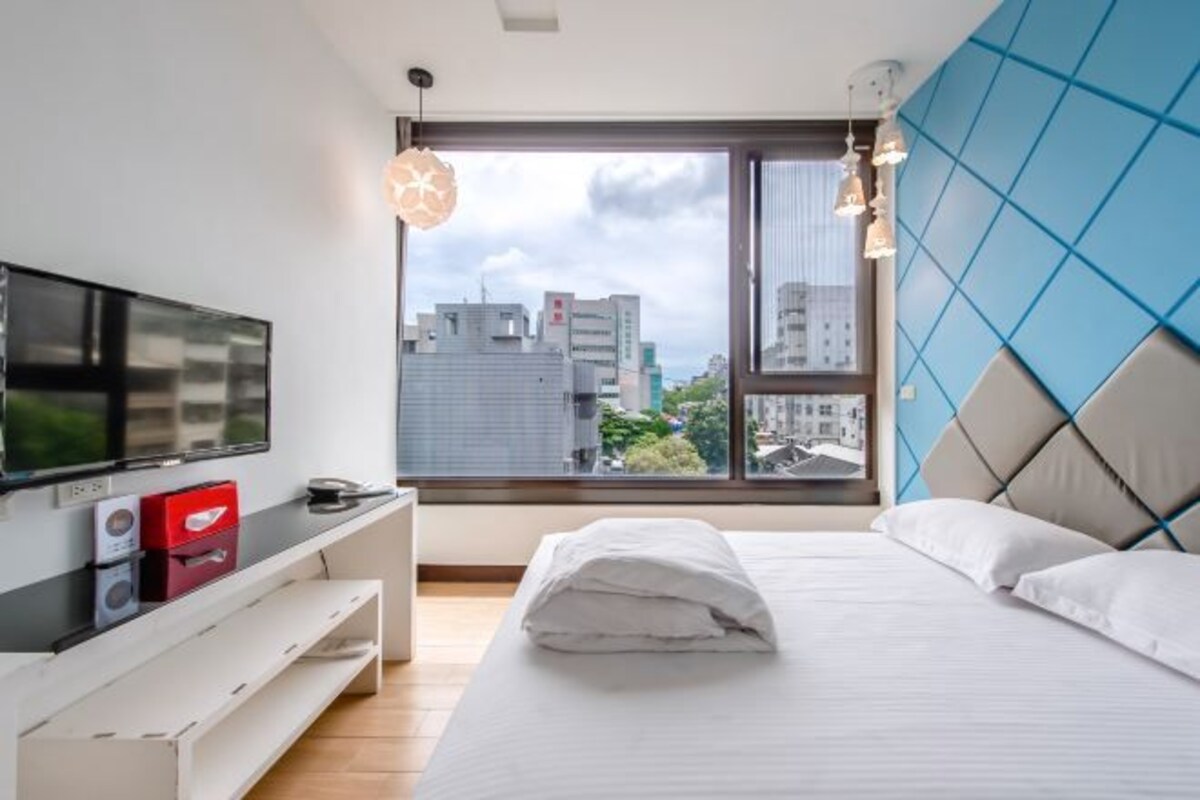 Habitació d'hotel moderna amb una finestra gran, un llit, una televisió i una taula consola blanca.