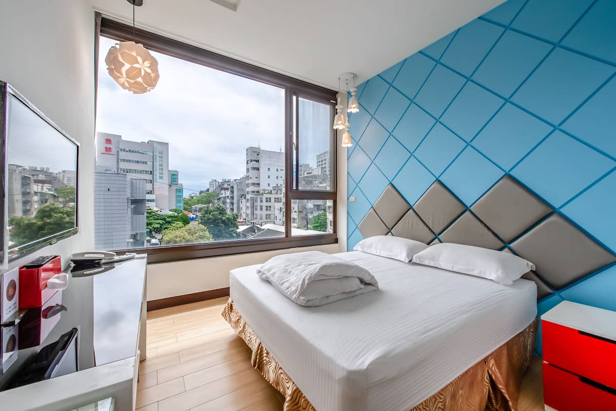 Un dormitorio moderno con un ventanal grande, una cama con sábanas blancas, una mesita de noche roja y una pared de acento azul.