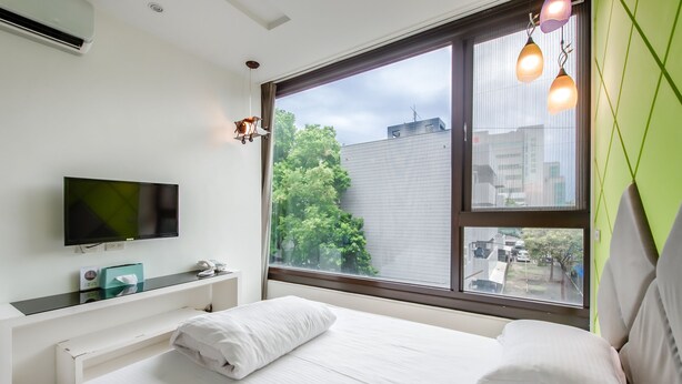 A modern hotel room with a large window, a flat-screen TV, a bedside table, and a bed with white linens. - Lightness Hotel (Tainan)