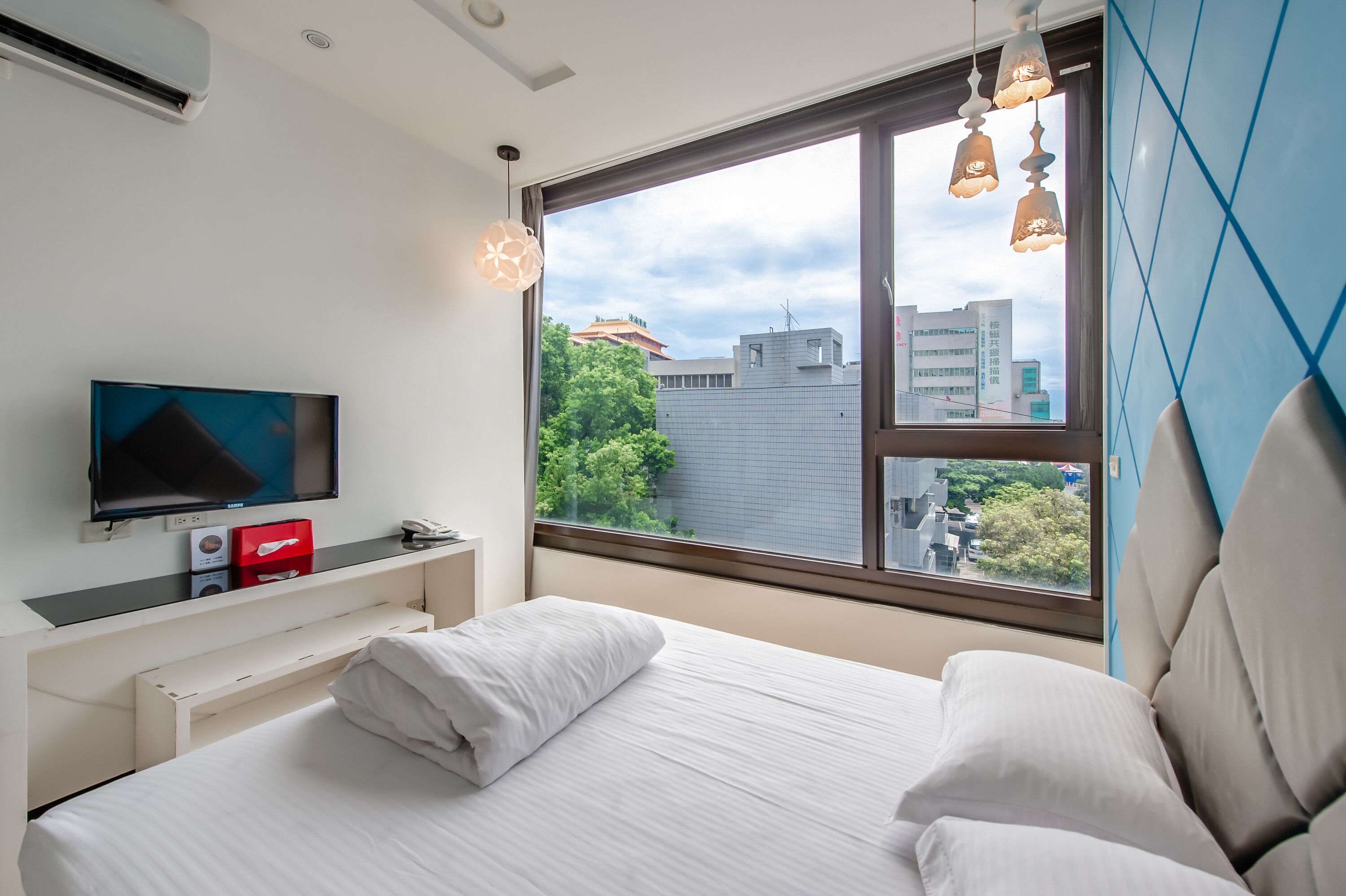 A modern hotel room with a large window, a flat-screen TV, a white bed, and a small desk with a red box and a phone.