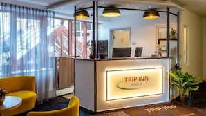 Uma área de recepção com o letreiro 'TRIP INN HOTELS', um balcão, duas cadeiras amarelas e uma escada ao fundo.