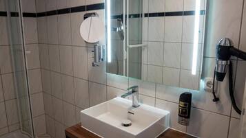 Un baño moderno con un lavamanos blanco, un cabezal de ducha de mano y un espejo con un espejo redondo encima.