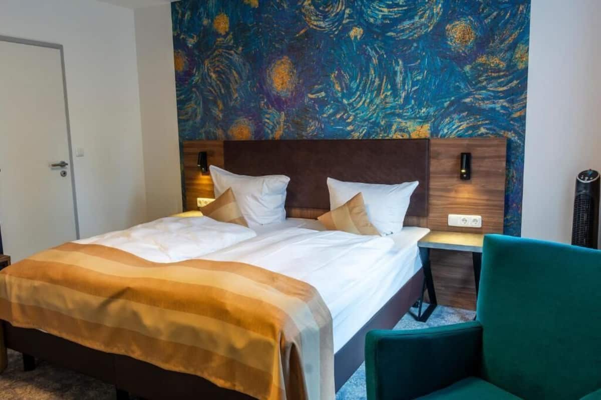 A hotel room with a bed, a nightstand, a green chair, and a wall with a painting.