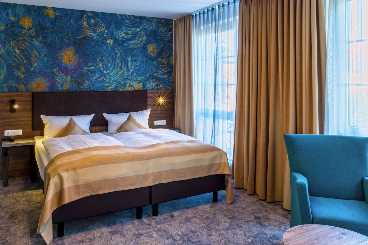 A hotel room with a large bed, a nightstand, a chair, and a wall with a blue abstract painting.