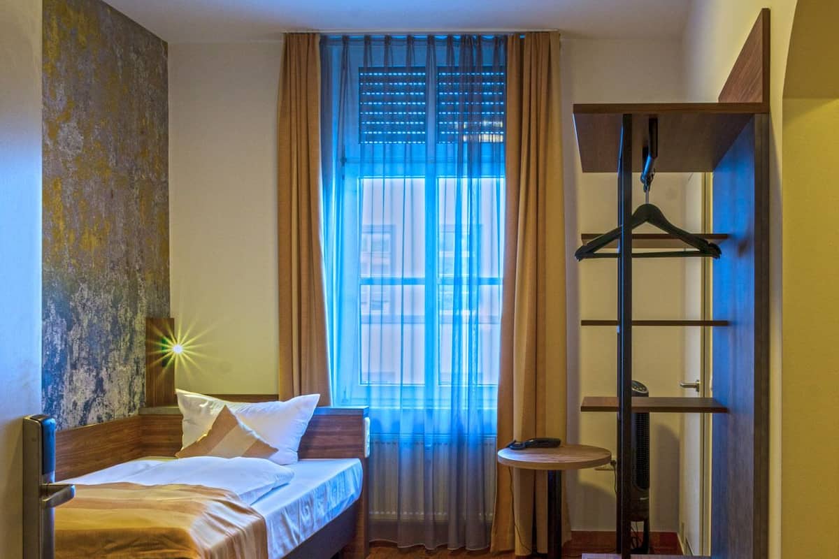 A hotel room with a bed, a small table, a hanging rack, and a view of the outside through a window with curtains.