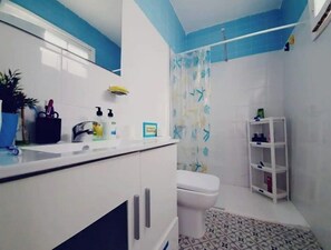 Casa de banho compacta com sanita, chuveiro com cortina, lavatório com artigos de higiene e uma pequena prateleira com frascos.