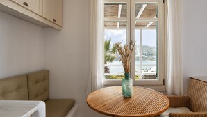A room with a round wooden table, a cushioned bench, and a view of the sea through a window with wooden shutters. - Villa Hola Mykonos (Mykonos)