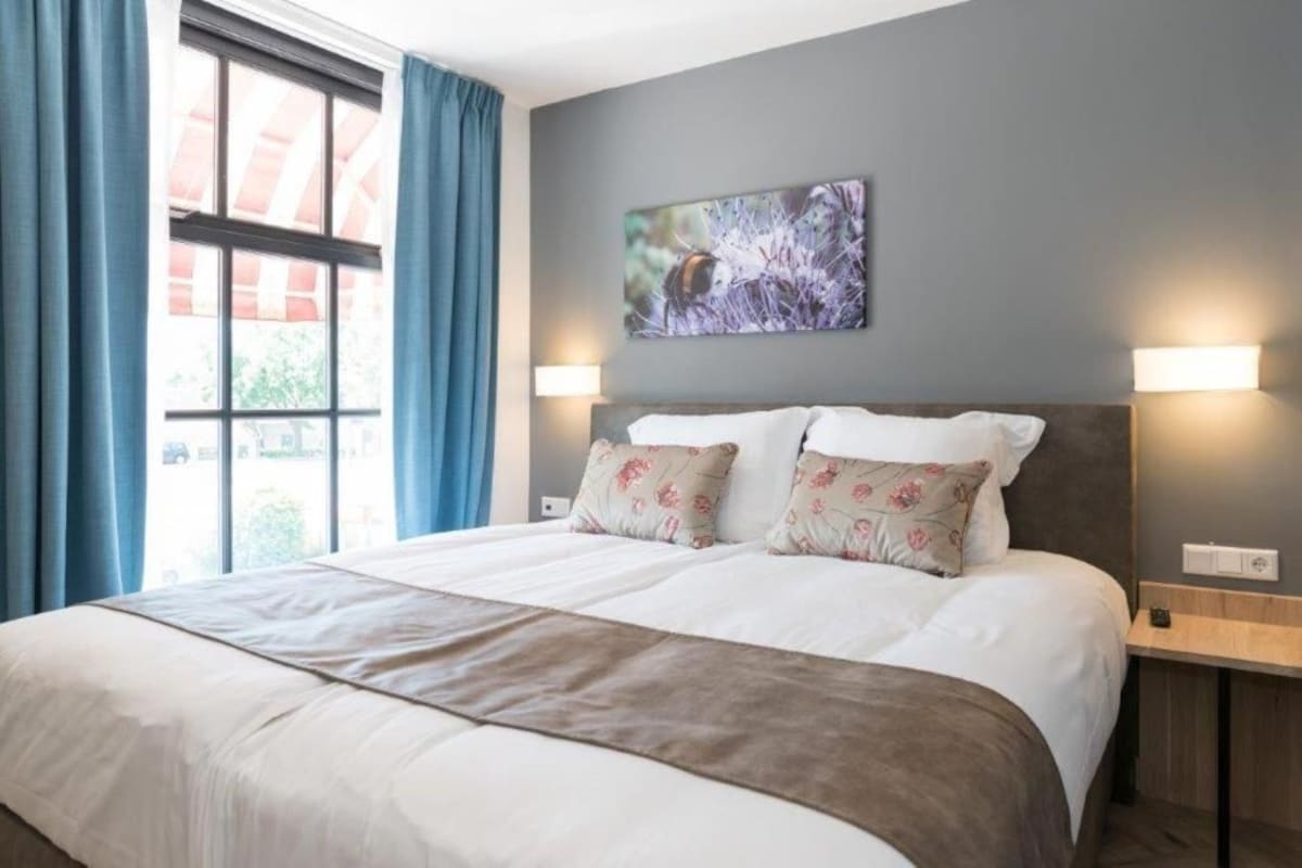 A hotel room with a large bed, two bedside lamps, a window with curtains, and a wall-mounted picture.