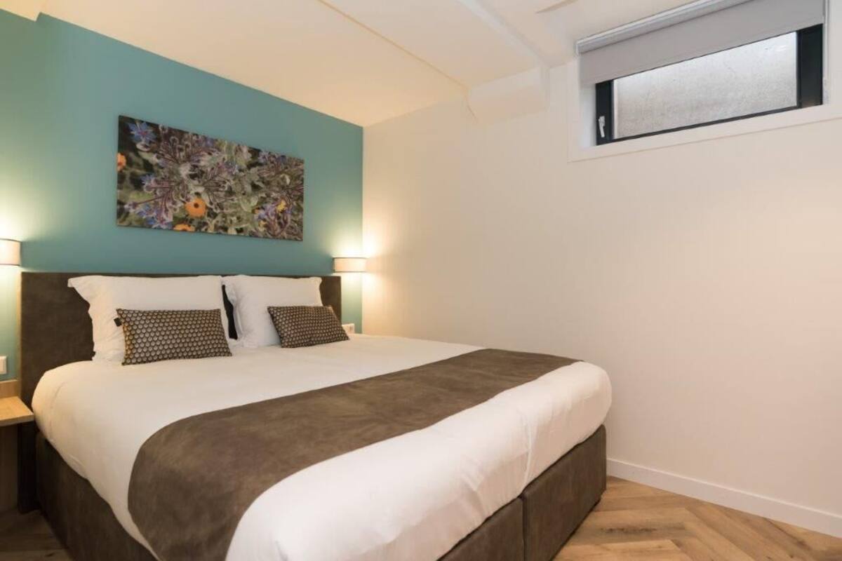 A bedroom with a bed, two bedside lamps, a floral artwork on the wall, and a window above the bed.