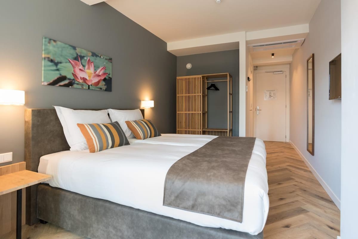 A hotel room with a large bed, bedside tables, a wall-mounted lamp, a wooden wardrobe, and a door leading to another room.