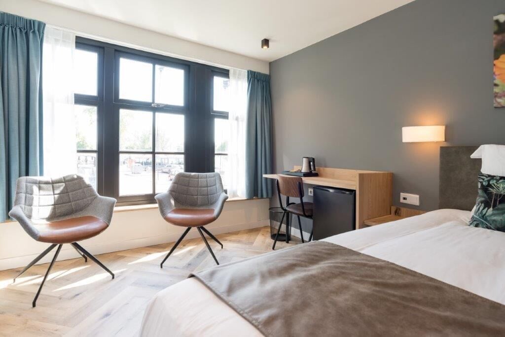 Une chambre d’hôtel avec un lit, deux chaises, un bureau et une fenêtre avec des rideaux.