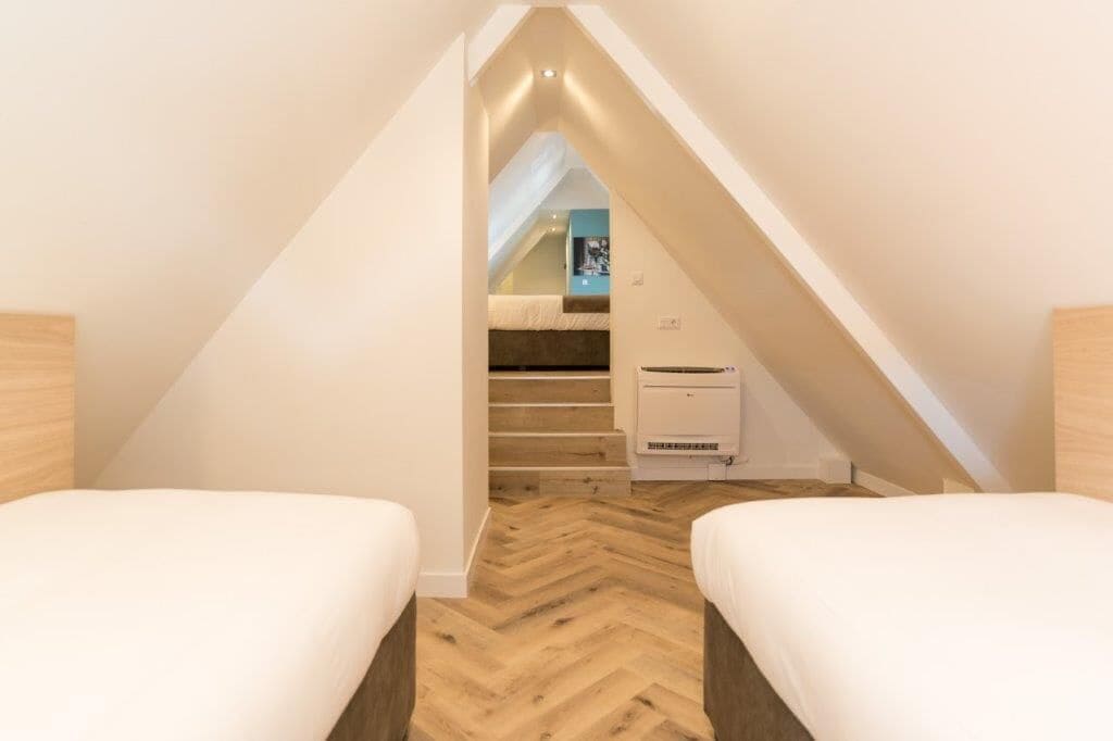 Un dormitorio en ático con espacio reducido, suelo de madera tipo espiga, dos camas, una escalera que lleva a una buhardilla y un equipo de aire acondicionado blanco.
