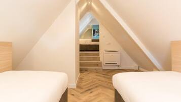 Un dormitorio en ático con espacio reducido, suelo de madera tipo espiga, dos camas, una escalera que lleva a una buhardilla y un equipo de aire acondicionado blanco.
