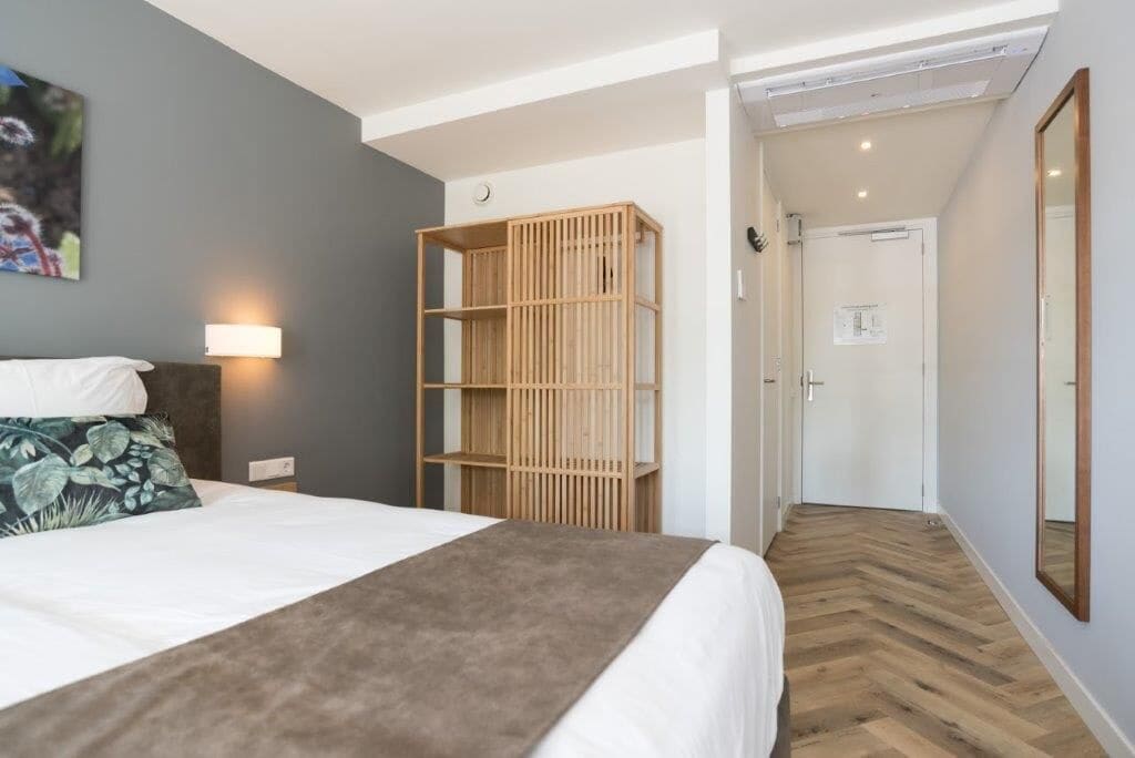 Een hotelkamer met een bed, een houten boekenkast, een wandlamp en een spiegel.