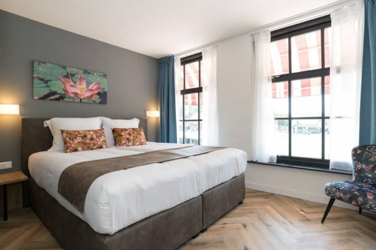 A bedroom with a large bed, two bedside lamps, a floral painting on the wall, and a window with curtains.