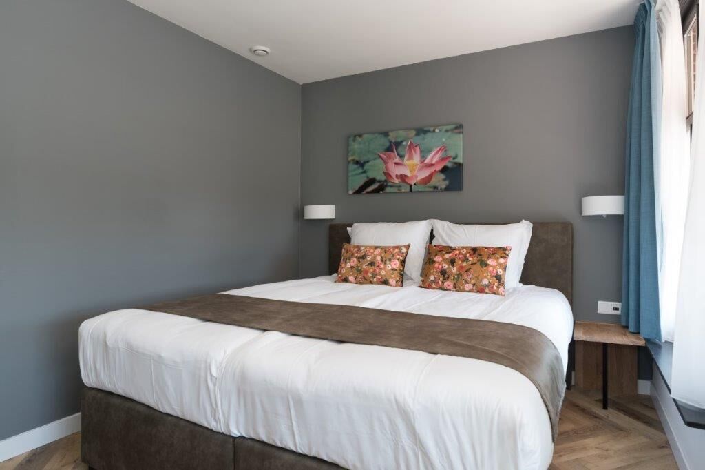 A bedroom with a large bed, a nightstand, a wall-mounted lamp, and a painting of a lotus flower.