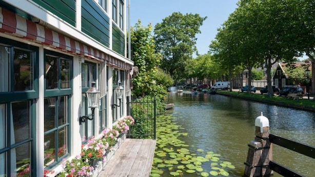 Un edificio junto a un canal, con una terraza de madera y jardineras florales, un estrecho canal con embarcaciones, y una fila de coches estacionados a lo largo de la calle.