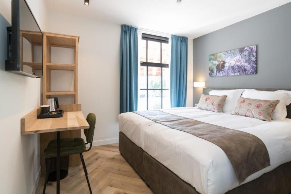 A hotel room with a bed, a desk with a chair, a window with curtains, and a wall-mounted artwork.
