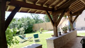 Ein möblierter Außenbereich mit Holz-Pergola, Steinsitzgelegenheiten und Blick auf einen Rasen mit Bäumen und einem Holzzaun.
