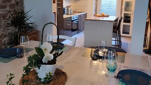 A dining area with a table set for a meal, a kitchen in the background, and a stone wall. - House 6 pers beach 3km town center 5mn walk (Crozon)