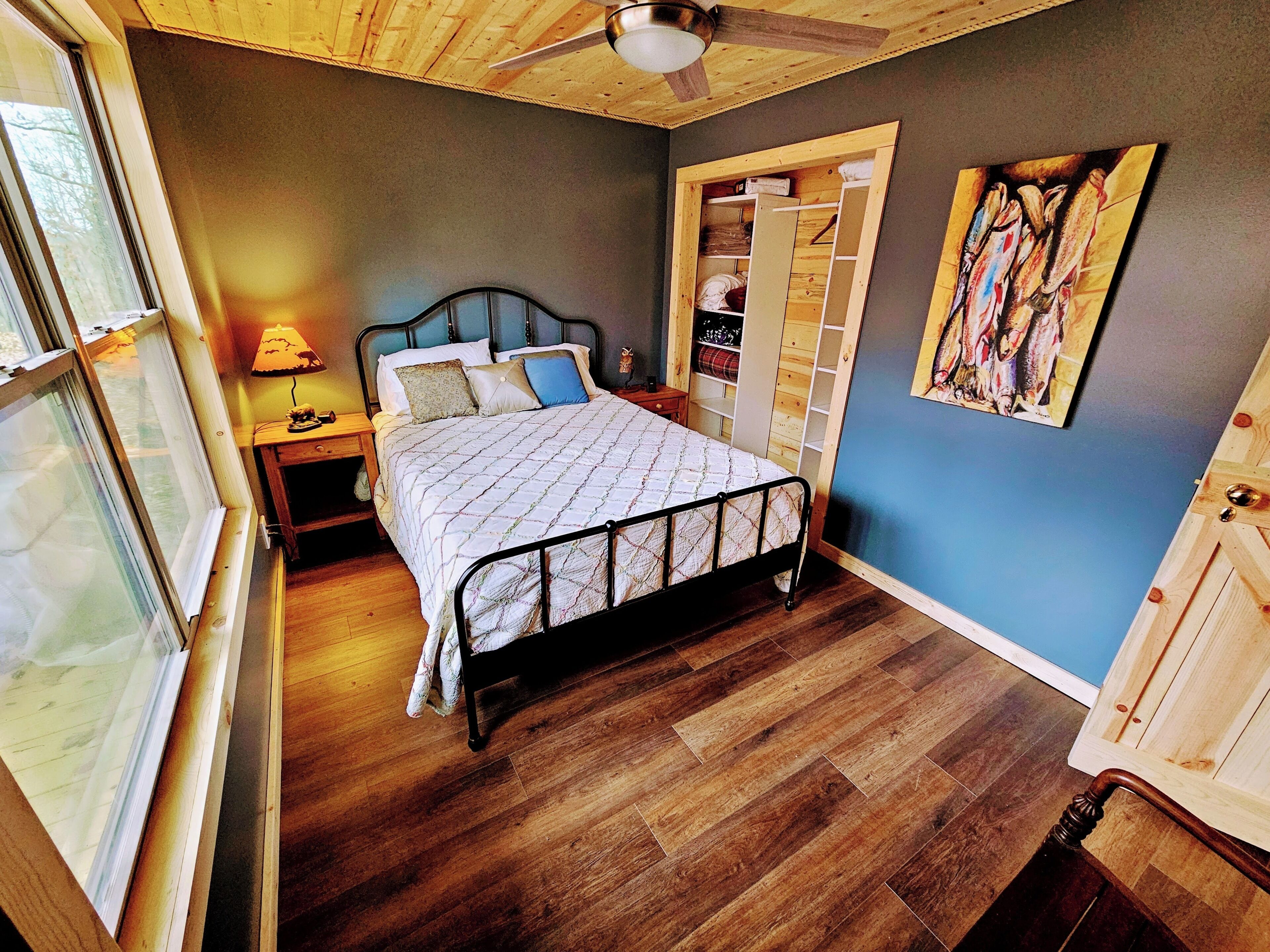 A bedroom with a bed, wooden floor, a ceiling fan, a nightstand, a window, and a wardrobe.